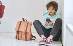 A young teen with a backpack beside him sits on the floor in a corner looking at a mobile phone.