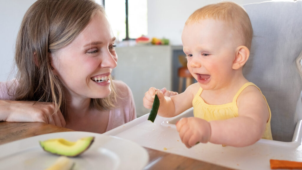 A happy mom watches her baby try eating solid foods with avocado slices.