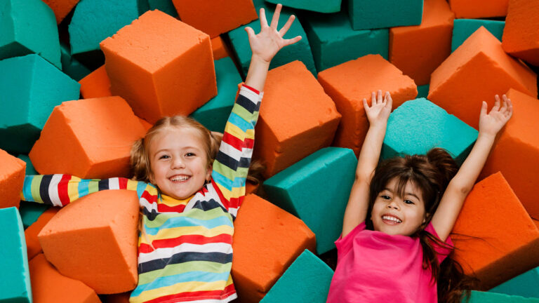 Two young girls play among orange and green foam blocks in an indoor kids playground.