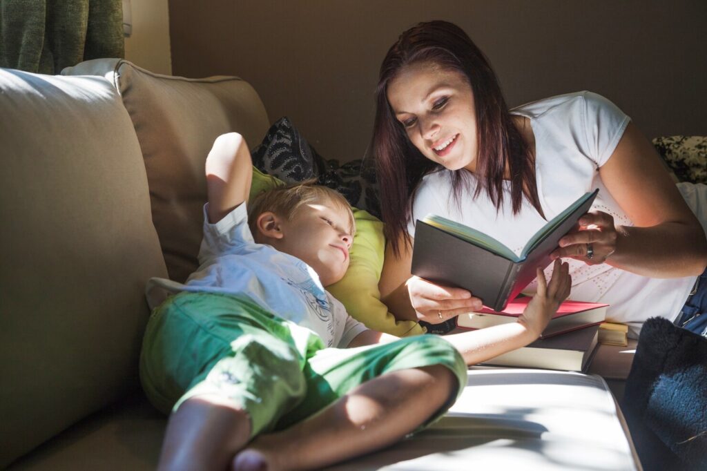 A mother and her young child sit on a cozy couch, under a blanket, reading a book together.