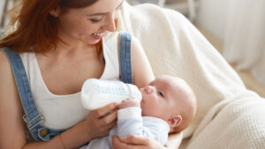 A smiling mother in denim overalls feeds her baby with a bottle.